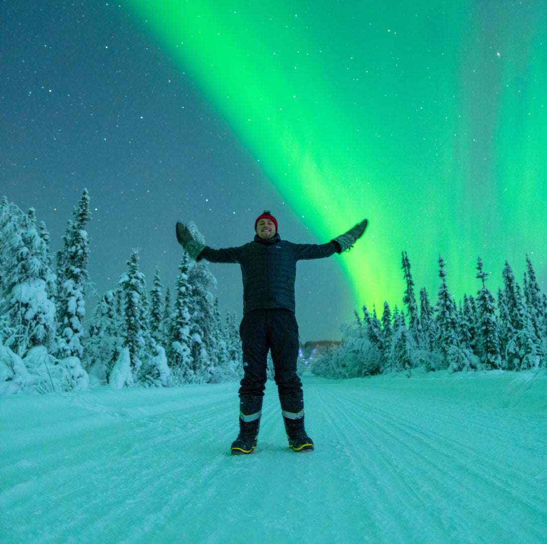 Vincent Ledvina standing in front of the aurora borealis on a winter's night on a road cutting through a snowy forest.