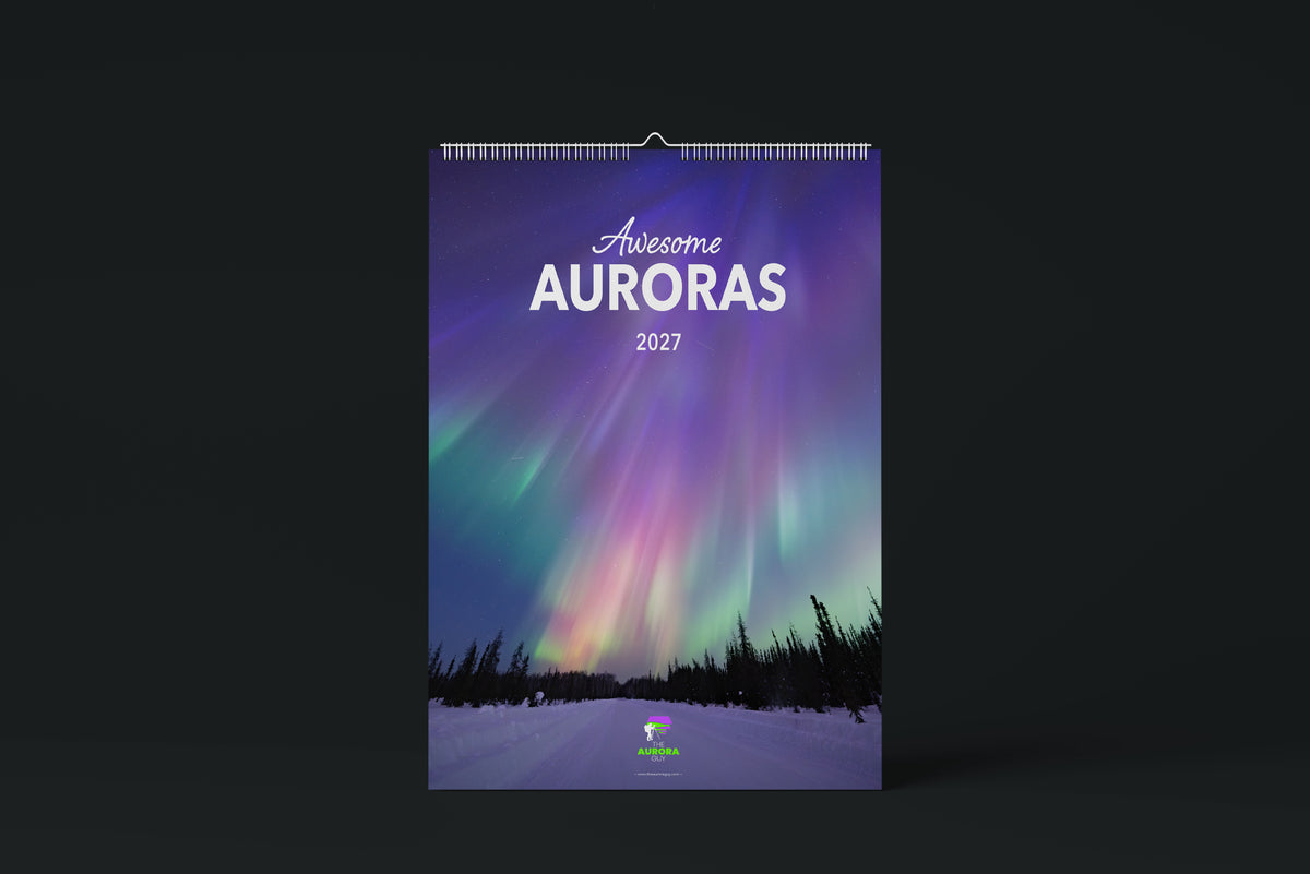 The 2027 Awesome Auroras wall calendar, its cover a photo of purple, pink and green aurora rays over spruce trees and a snowy road, shown against a dark background.