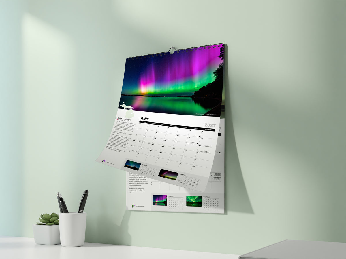 The 2027 Awesome Auroras calendar hanging on a pale green wall, open to June: "Rainbow Magic" by Lisa Lirones, pink and green aurora reflected in Torch Lake, Michigan.