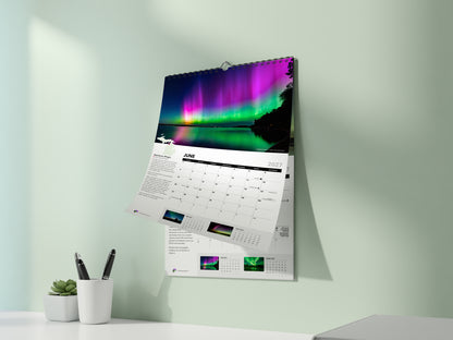 The 2027 Awesome Auroras calendar hanging on a pale green wall, open to June: "Rainbow Magic" by Lisa Lirones, pink and green aurora reflected in Torch Lake, Michigan.