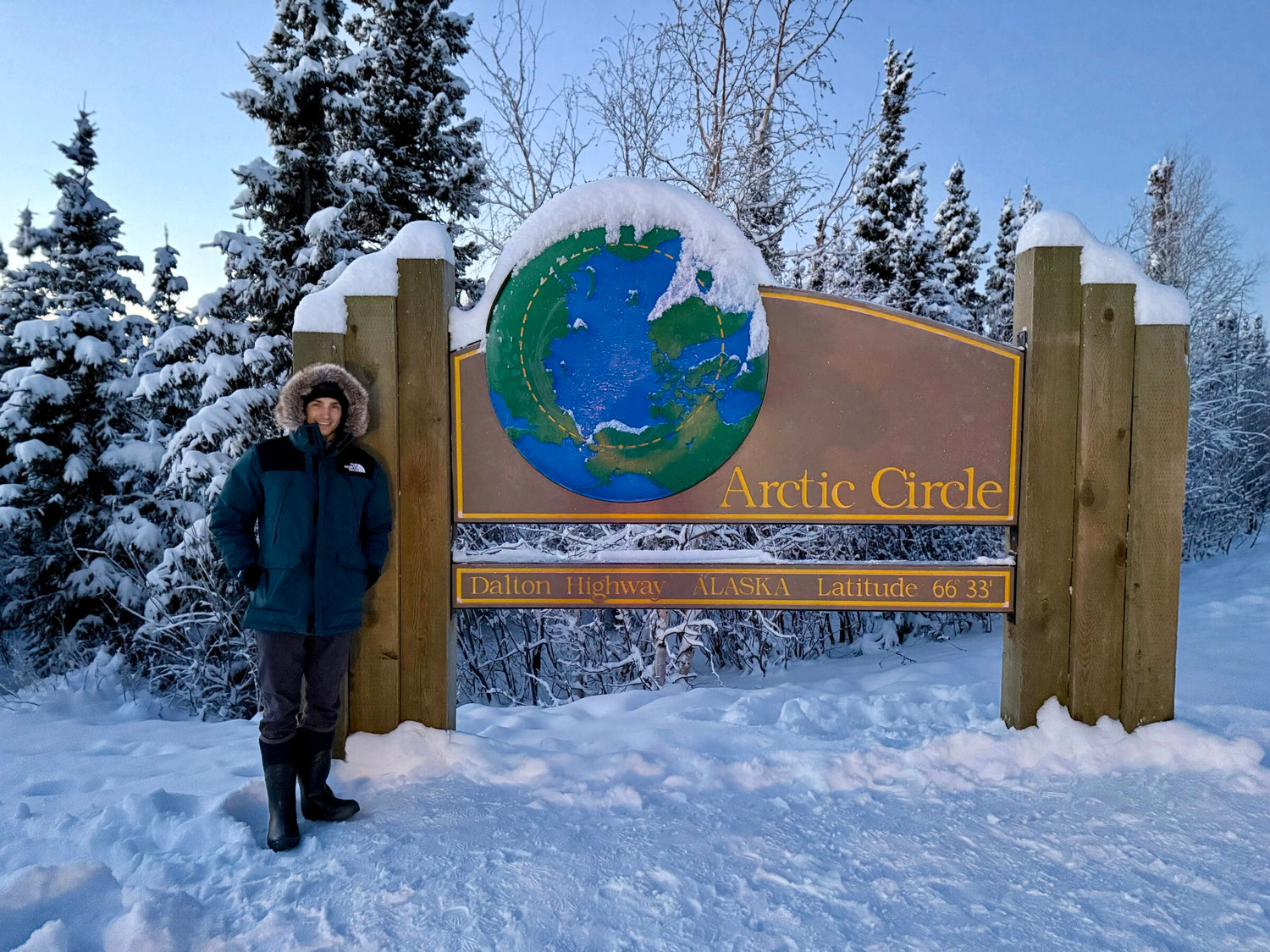 Guest standing at the Arctic Circle sign on the Dalton Highway in Alaska at latitude 66 degrees 33 minutes north, on a Midnight Sky Adventures tour
